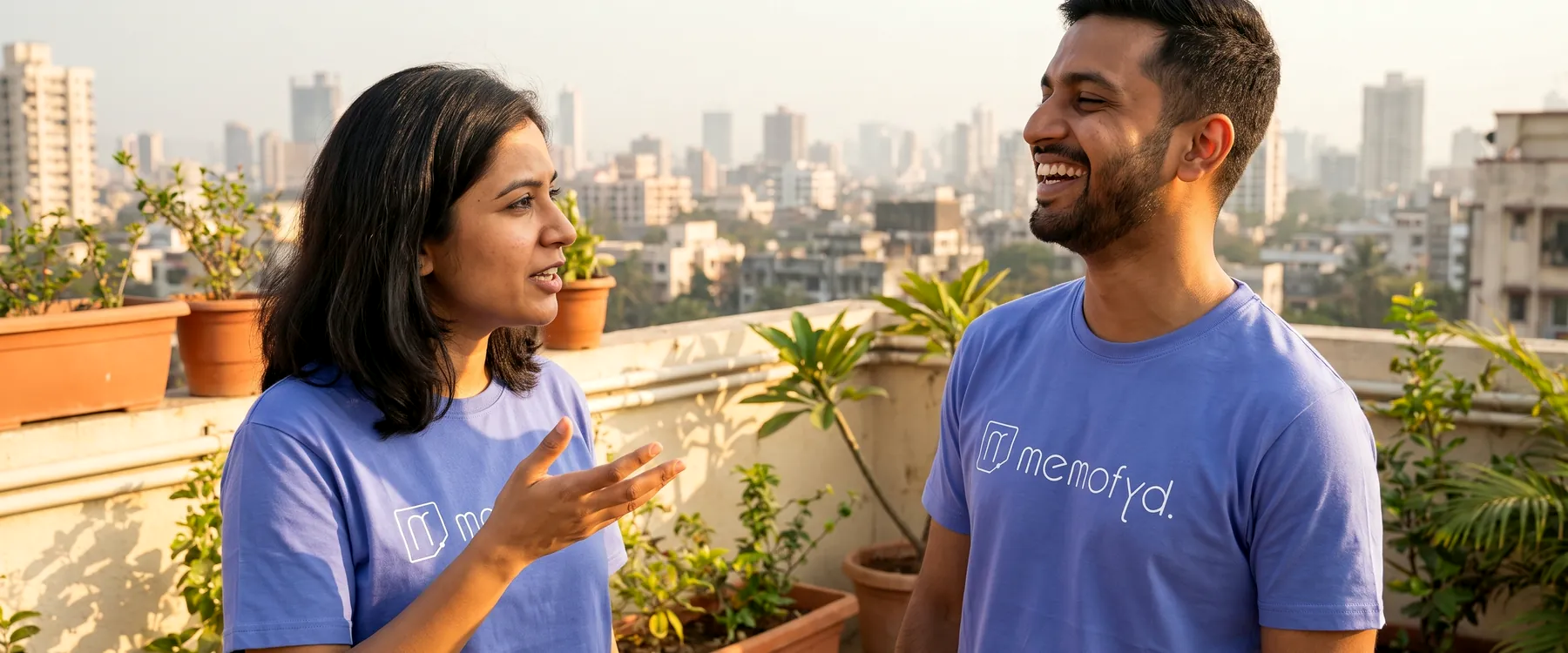 Two colleagues in Memofyd t-shirts talking on a rooftop terrace above an Indian city at golden hour.